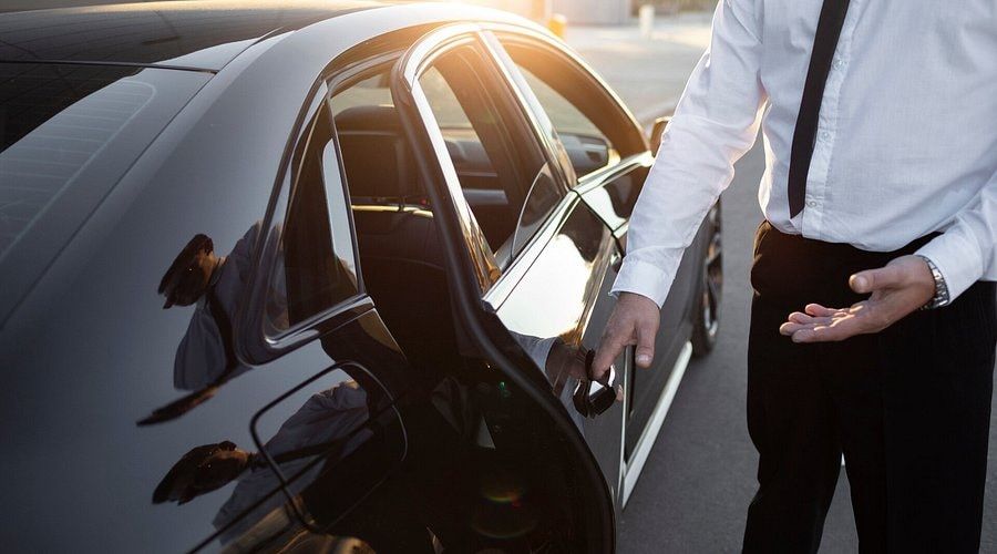 Chauffeur en chemise blanche ouvrant la portière arrière d’une berline noire au soleil couchant